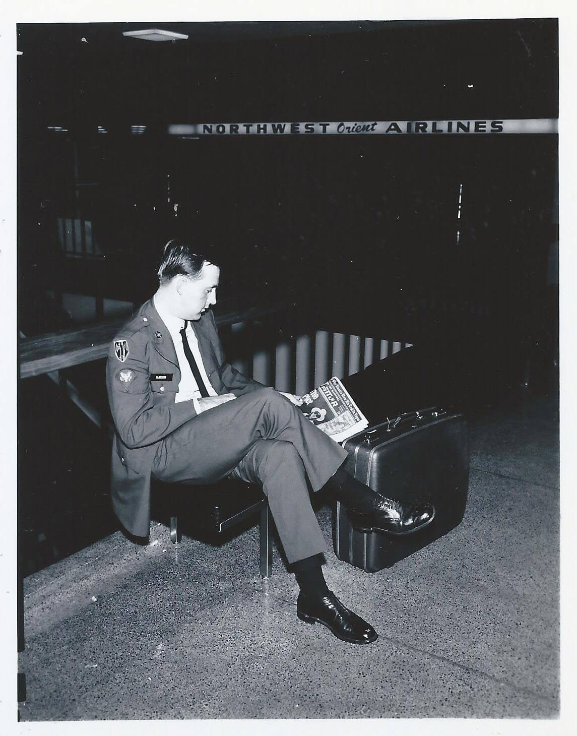 A soldier in his dress blues sits in an airport with his luggage, reading a newspaper.