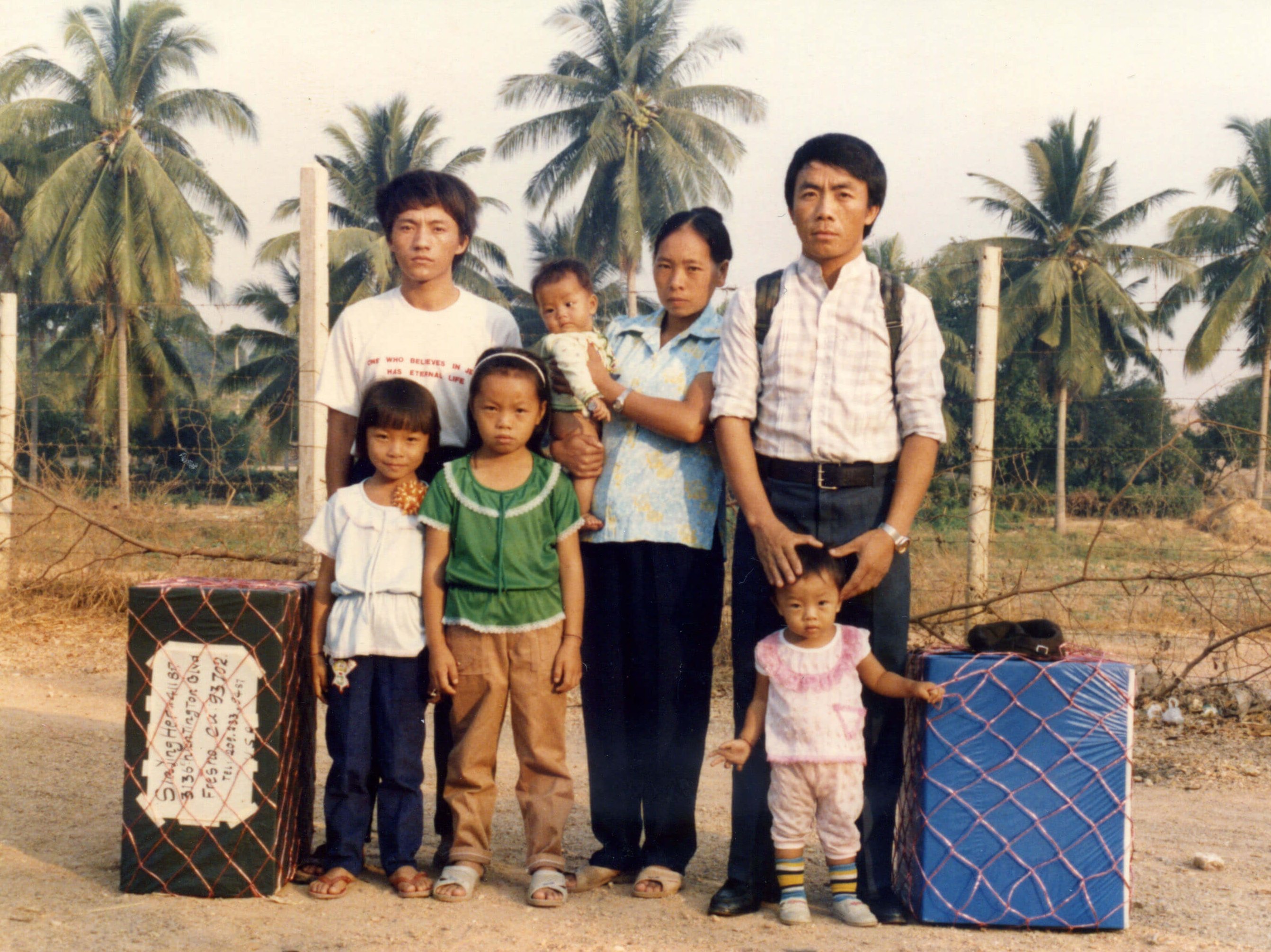 Portrait of a Hmong family of 7 standing outside in a tropical climate, luggage stacked on either side of them.