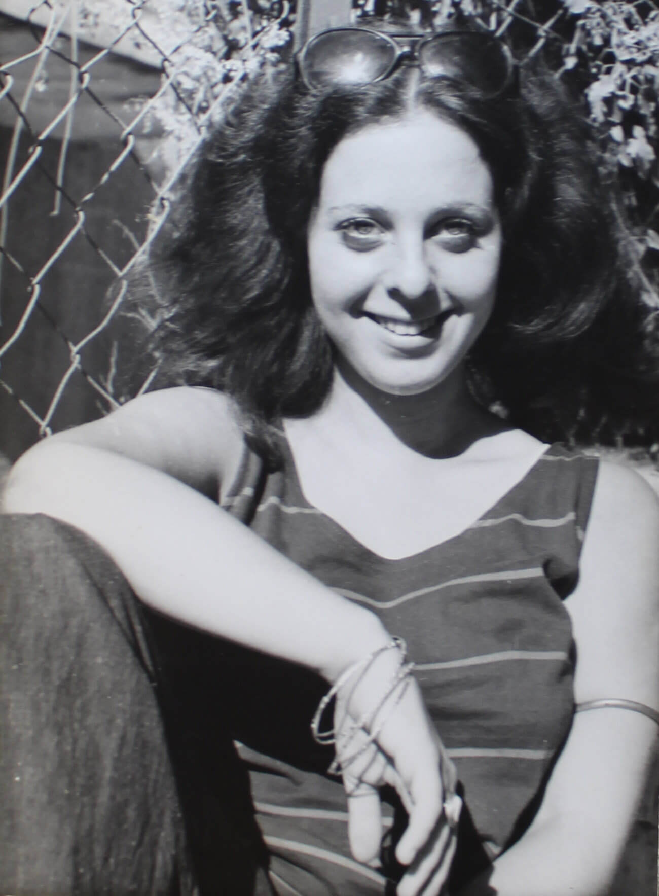 Photo of a young woman casually sitting against a chain-link fence in the summer.