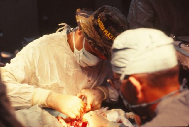 A couple men operating on a person; all are wearing scrubs; one of the surgeons is wearing a camouflage boonie hat.