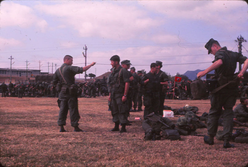 Several soldiers dressed in green uniforms putting on equipment, figure on left has arm up and pointing finger either at someone or something off to the right.