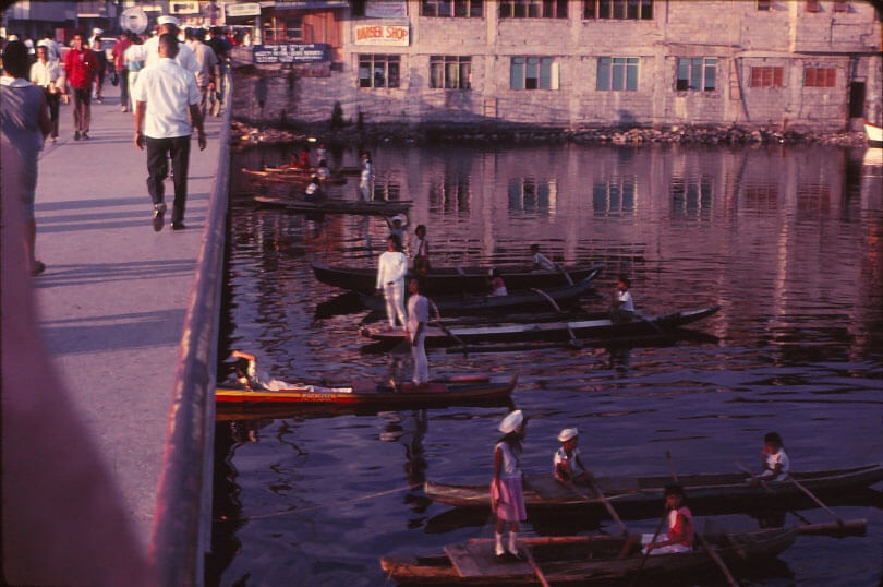 A concrete pier with pedestrians walking alongside the water, where a number of canoes with people.