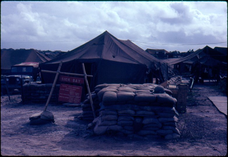 Red sign with "Sick Bay" on it; canvas tents with sandbag walls surrounding them.
