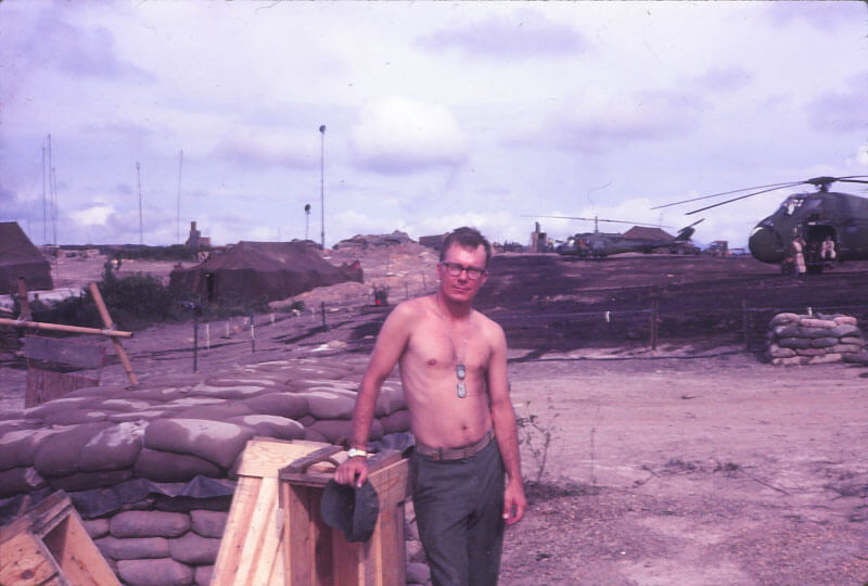 Young shirtless man, standing with his right hand on a sandbag wall. Behind him is the military base with two helicopters.