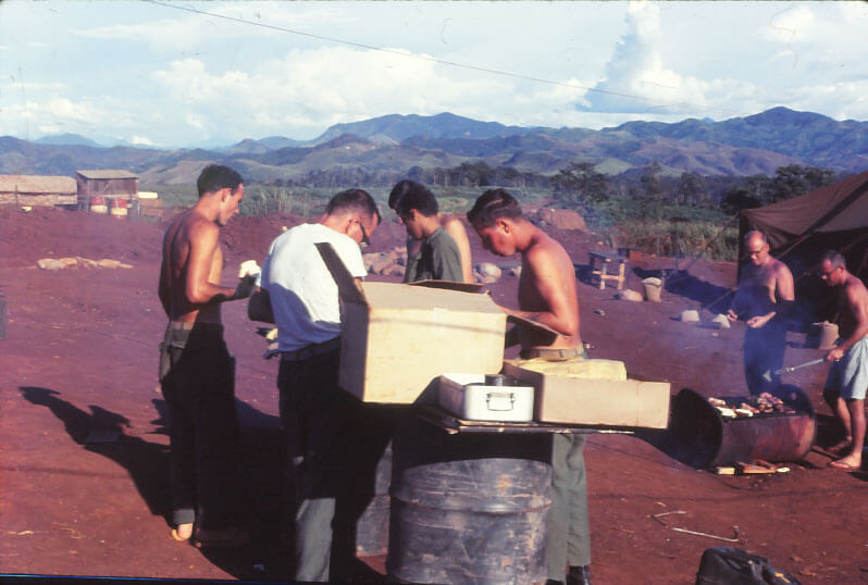 Five soldiers standing around a makeshift table resting on barrel legs, two men stand in the background over a grill that was cut out of a barrel.