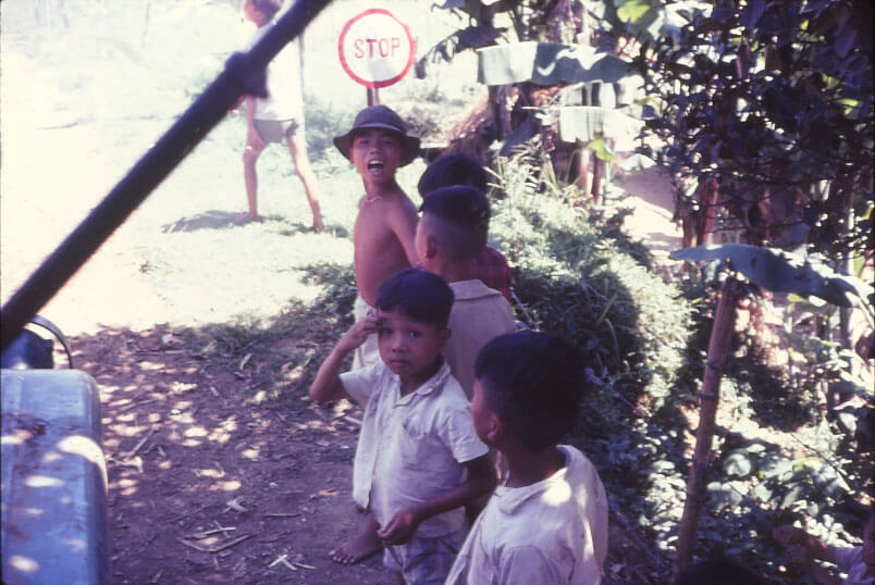A small group of Vietnamese children, one appears to be saluting, one appears to be yelling at the photographer, who seems to be in a truck.