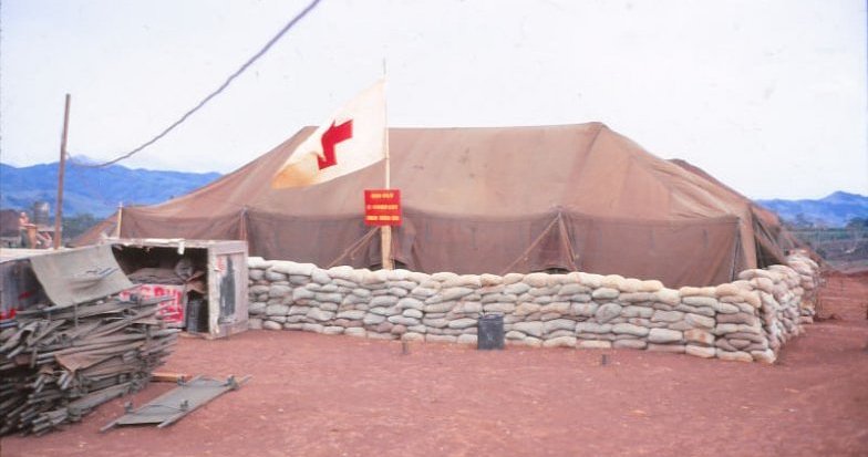 Hospital tent with a red cross flag flying outside; there is pile of cots outside the tent, sandbag wall surrounds the tent.