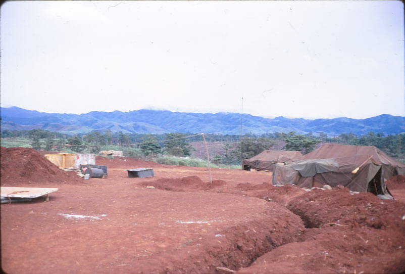 A trench with some canvas tents; hills and valleys and trees in the background.