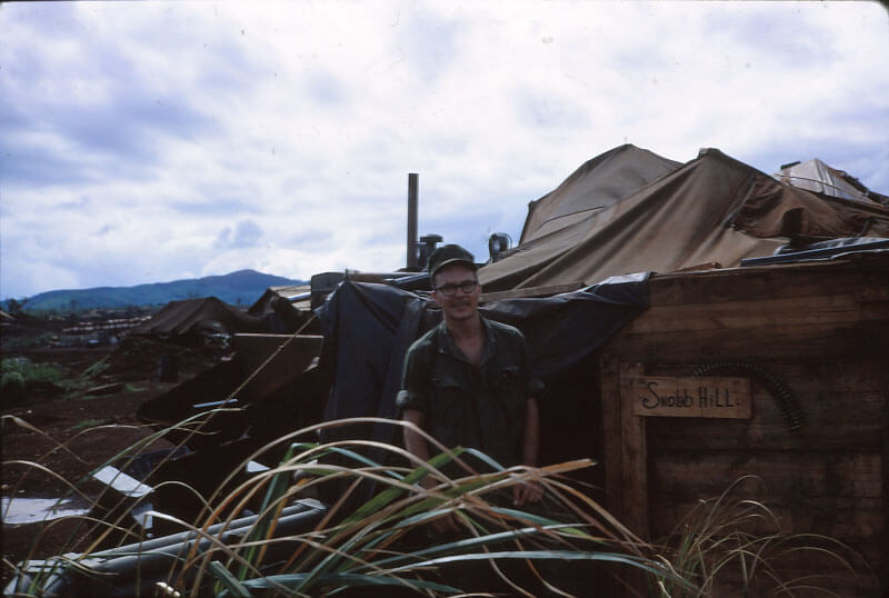 Soldier wearing green fatigues standing outside of a quarters consisting of a wood and a canvas roof; sign on the door "Snob Hill".