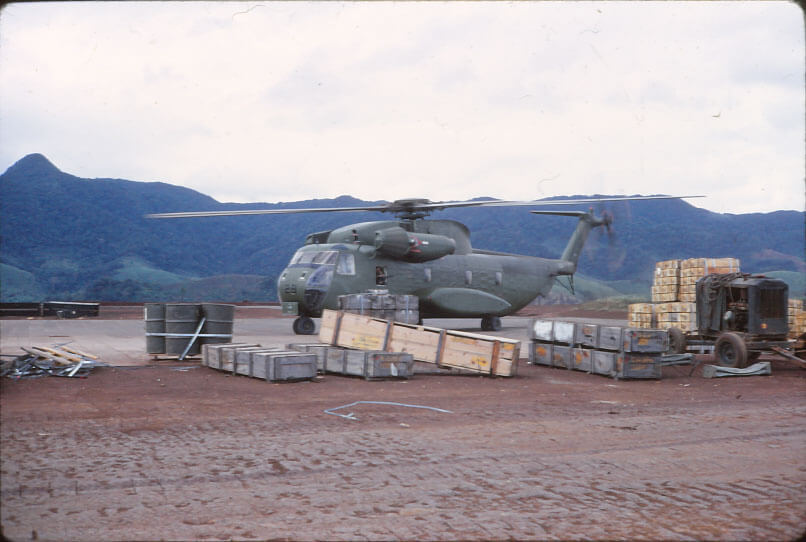 Helicopter sitting on the pad with boxes of equipment and supplies, some barrels and an engine or machine on a trailer.