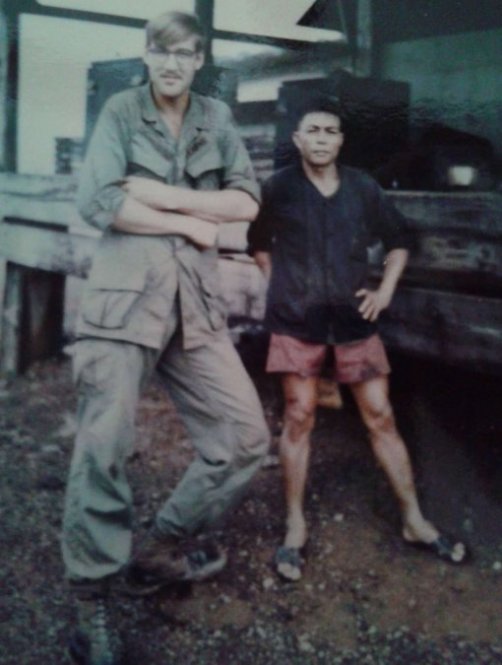 A young, tall soldier in uniform, standing next to a diminutive Asian man wearing shorts and sandals.