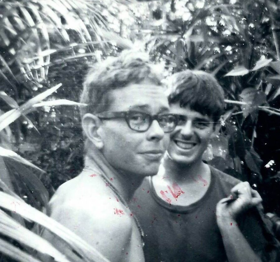 Two young men--one shirtless and with glasses, the other in a dark t-shirt--in the jungle.