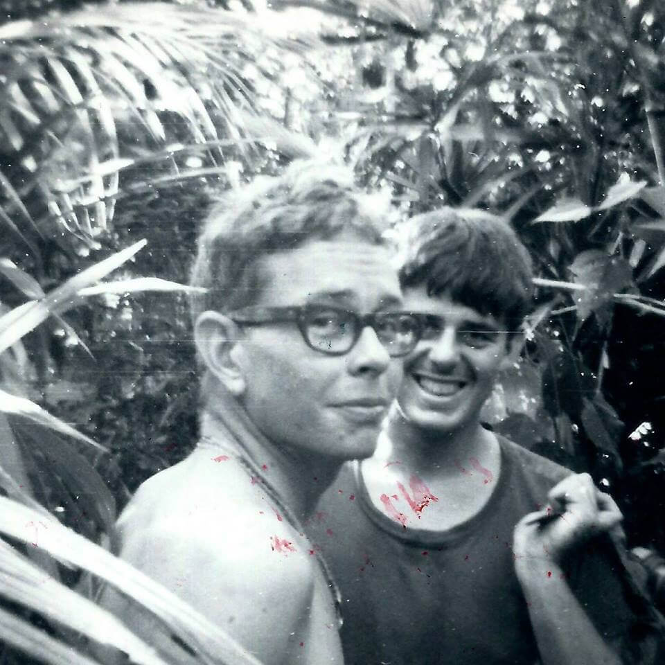 Two young men--one shirtless and with glasses, the other in a dark t-shirt--in the jungle.