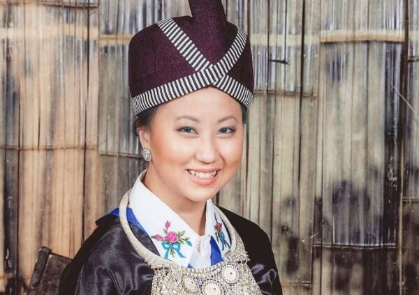 A young Hmong woman in traditional Hmong costume, in front of a bamboo backdrop.