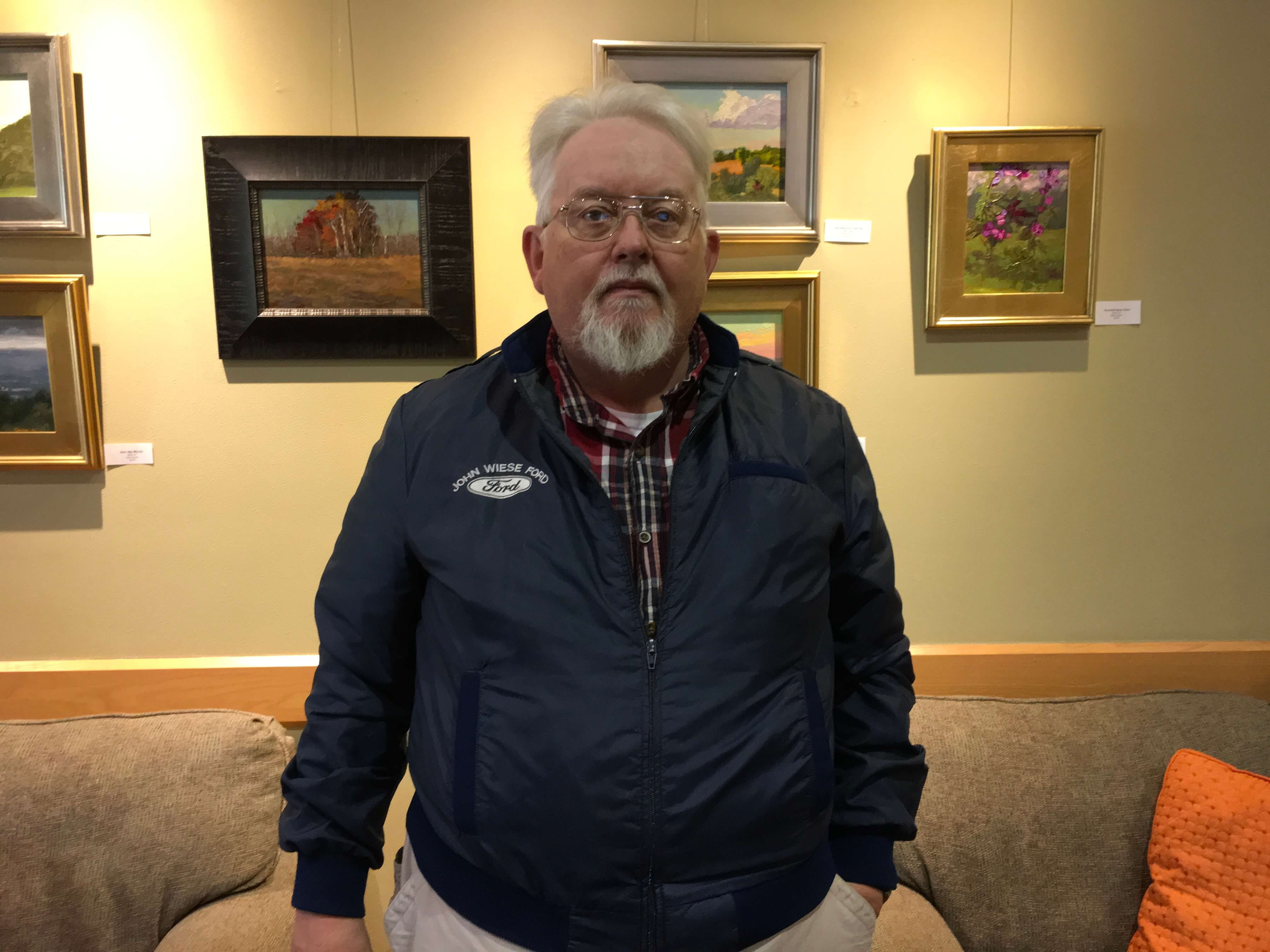 Man in jacket poses in front of picture frames on a wall.