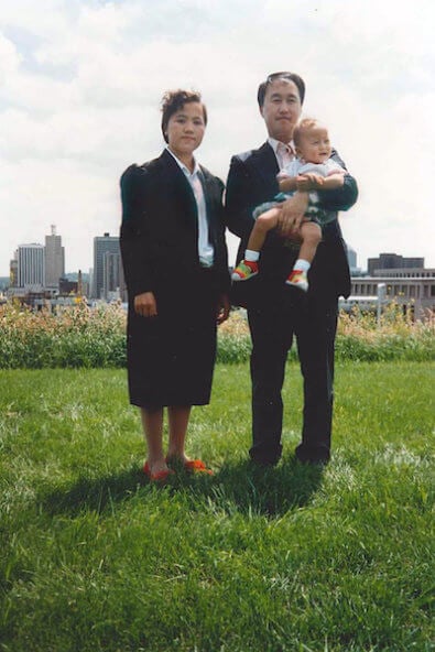 A Hmong couple and their young child stand on a grassy lawn, city buildings in the background.