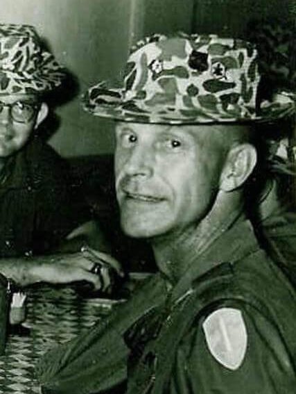 An older U.S. soldier seated at a table, looking casually over his left soldier toward the camera.