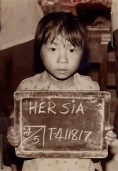 Refugee ID photo of a young Hmong girl holding a small chalk board that reads "HER SIA, 4/5, T-411817."
