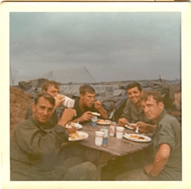 Young Vietnam soldiers sit at a table outside eating food.