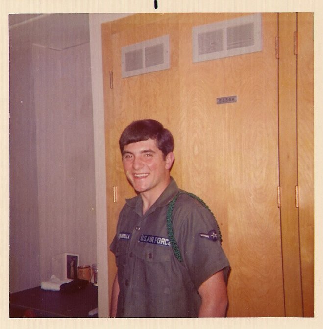 A smiling young man in a US Air Force uniform standing inside a building, in front of a wooden door.