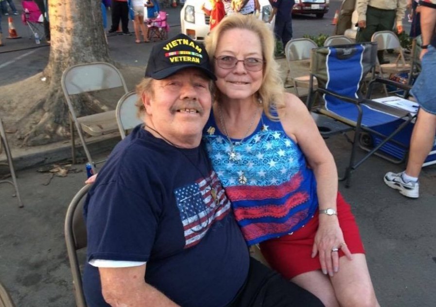 Contemporary photo of an older veteran and a woman, both wearing American flag apparel, seated outside at some sort of gathering.