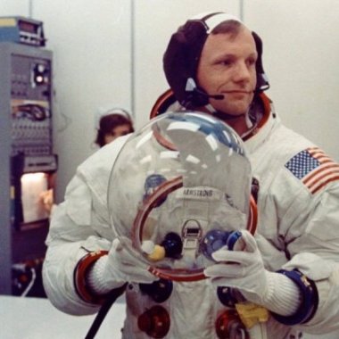Neil Armstrong in a white space suit, holding his helmet in his hands and glancing off to his left, in some sort of control room.