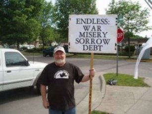 Contemporary photo of an older gentleman in a black Veterans for Peace t-shirt and ball cap, holding a sign that says "Endless War, Misery, Sorrow, Debt; Let's Try Peace".
