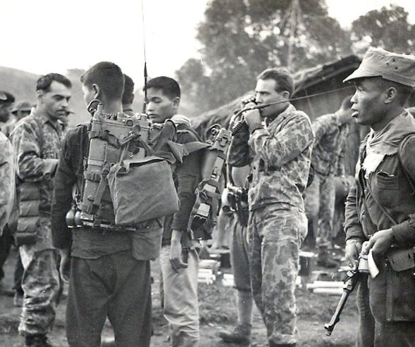 US and Asian soldiers with radios and rifles, standing outside an encampment.