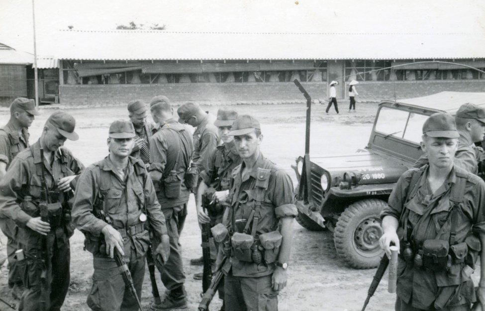 A group of U.S. soldiers and a Jeep on a street. Two Asian women walk on the other side of the street in the background.