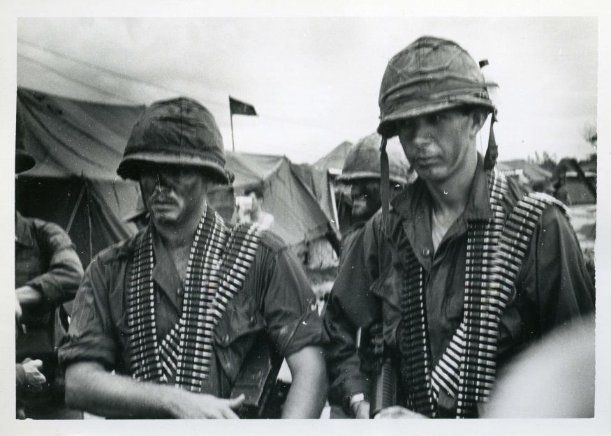 Close up of two US soldiers outside an encampment, wearing face paint and full gear including several bandoliers each draped on their shoulders.