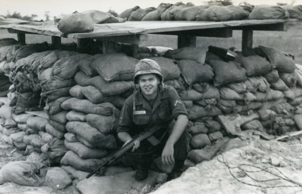A young US soldier squatting outside a sandbag bunker, rifle in hand.