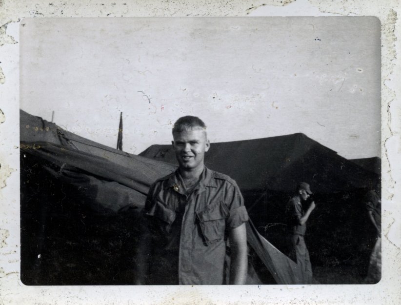 A weathered photo or slide of a young man in his Army shirt, standing outside an encampment.