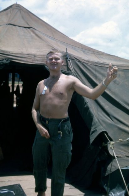 A shirtless US soldier with dog tags around his neck, standing outside a large canvas tent and zipping up his fly.