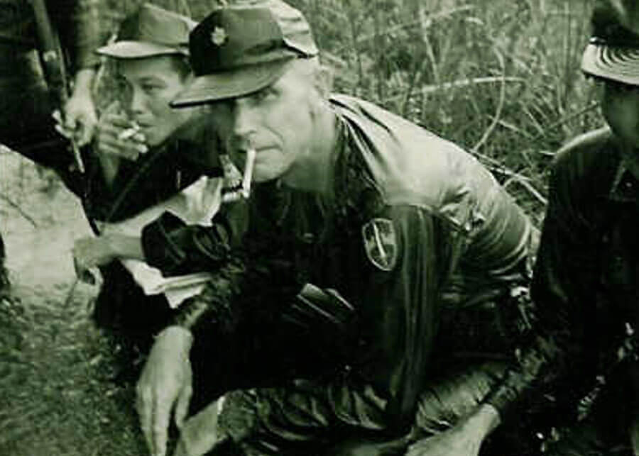 An older U.S. soldier crouched down with Asian soldiers, smoking a cigarette.