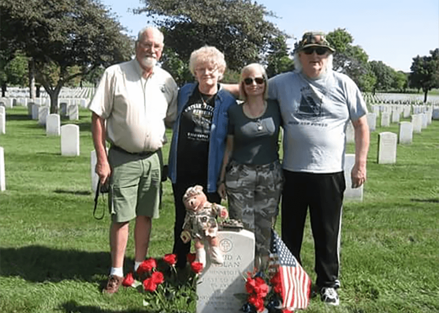 Contemporary photo of four people standing behind a white marble tombstone decorated with a miniature American flag, teddy bear in uniform, red carnations.