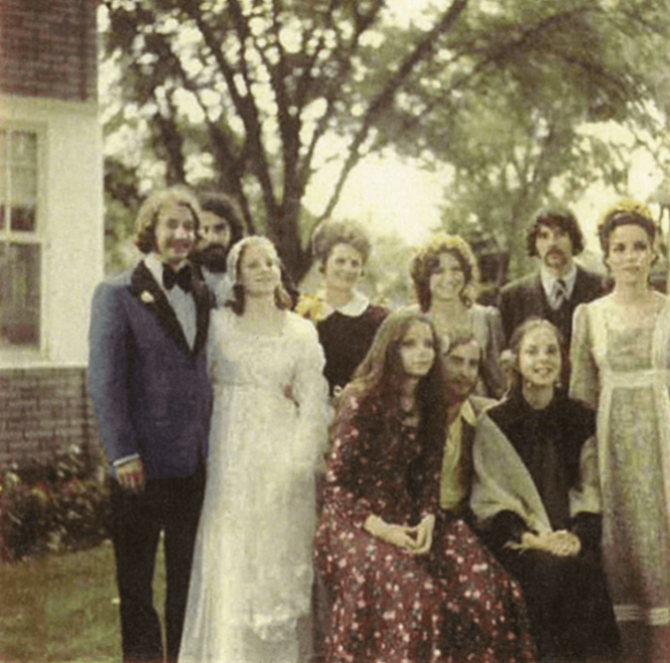 A family photo of 10 adults, 2 of which are a bride and groom. The photo is taken outside in a courtyard or yard.