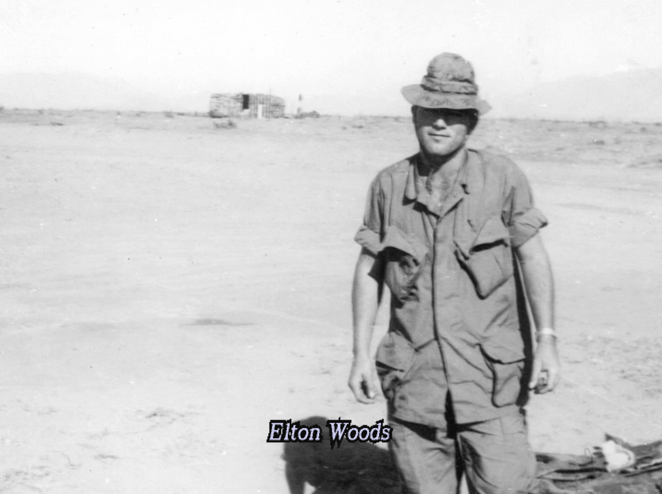 Photo of a soldier in uniform and a boonie hat, standing in a desolate landscape. Text on photo says "Elton Woods."