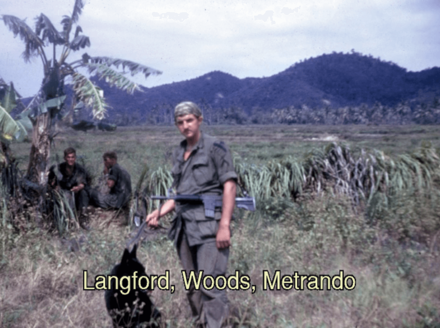 One soldier standing in the foreground, two soldiers crouching in the shade in the background. Grasses and mountains populate the landscape. Text on photo says "Langford, Woods, Metrando."
