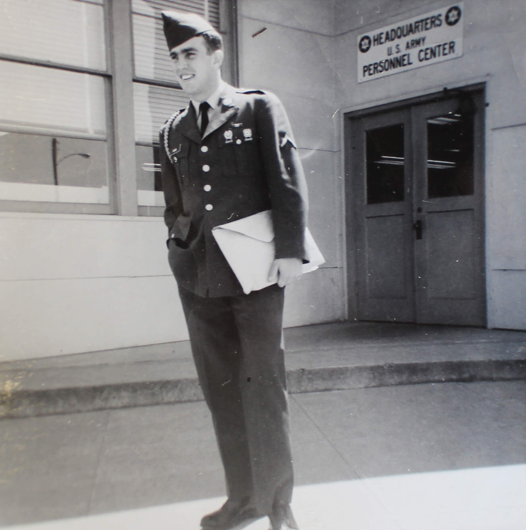 Black and white photo of a young soldier standing in front of the US Army Headquarters Personnel Center.