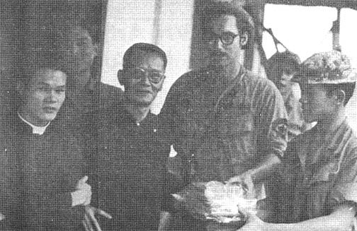 An Asian Catholic priest, another Asian man, a US soldier, and an Asian soldier pose for a photo. The two soldier hold a small package.