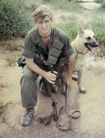 A young soldier kneeling in the dirt, a German shepherd sitting behind him.