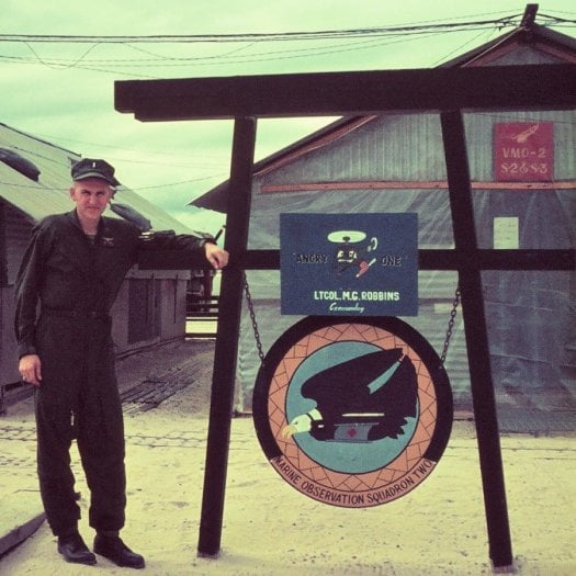 A young soldier standing in front of a hooch, leaning on a sign that says "Angry One LtCol M.G. Robbins Marine Observation Squadron Two".