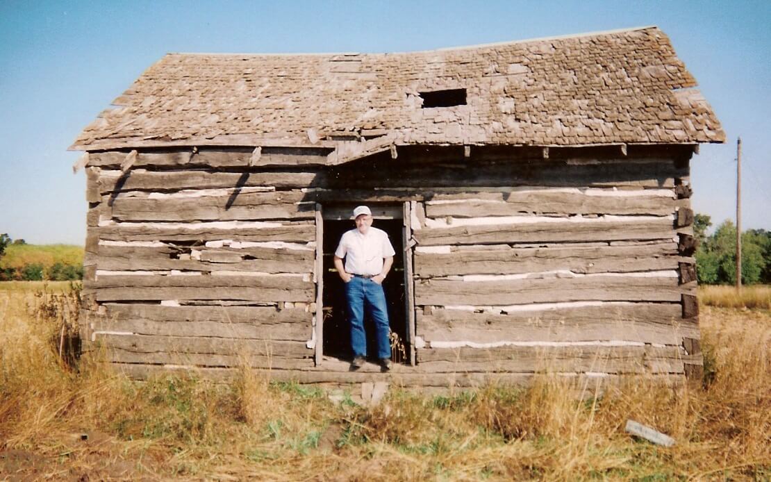 An older gentleman standing inside the doorway of a crude log building.