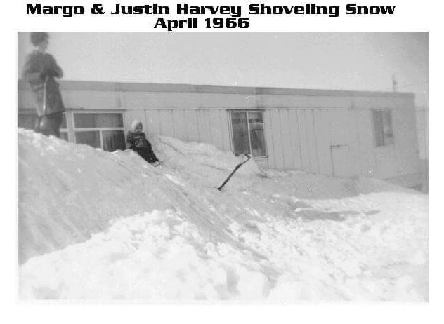 A woman and a child standing and playing on a very high mound of snow. The womans head is higher than the roofline of the single-story building she stands in front of.