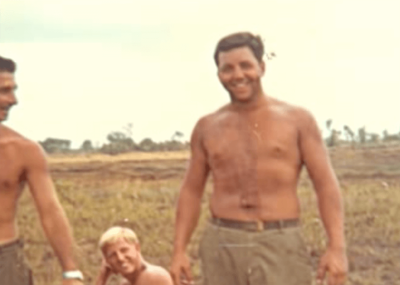 A shirtless soldier with a wide grin on his face; two other smiling soldiers are in the background.