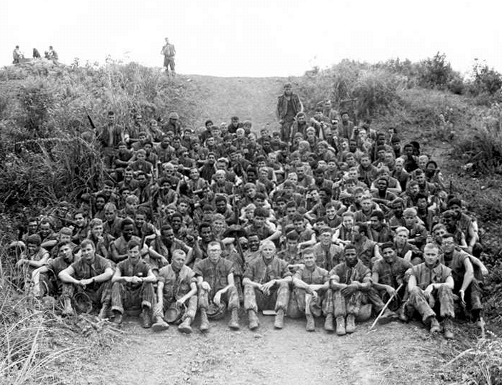 A large company of US soldiers sitting on a hill, posing for a photo.