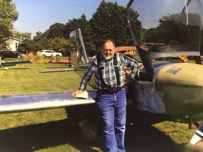 Contemporary photo of an older gentleman in jeans, a plaid shirt, and suspenders, leaning against the wing of a small plane.