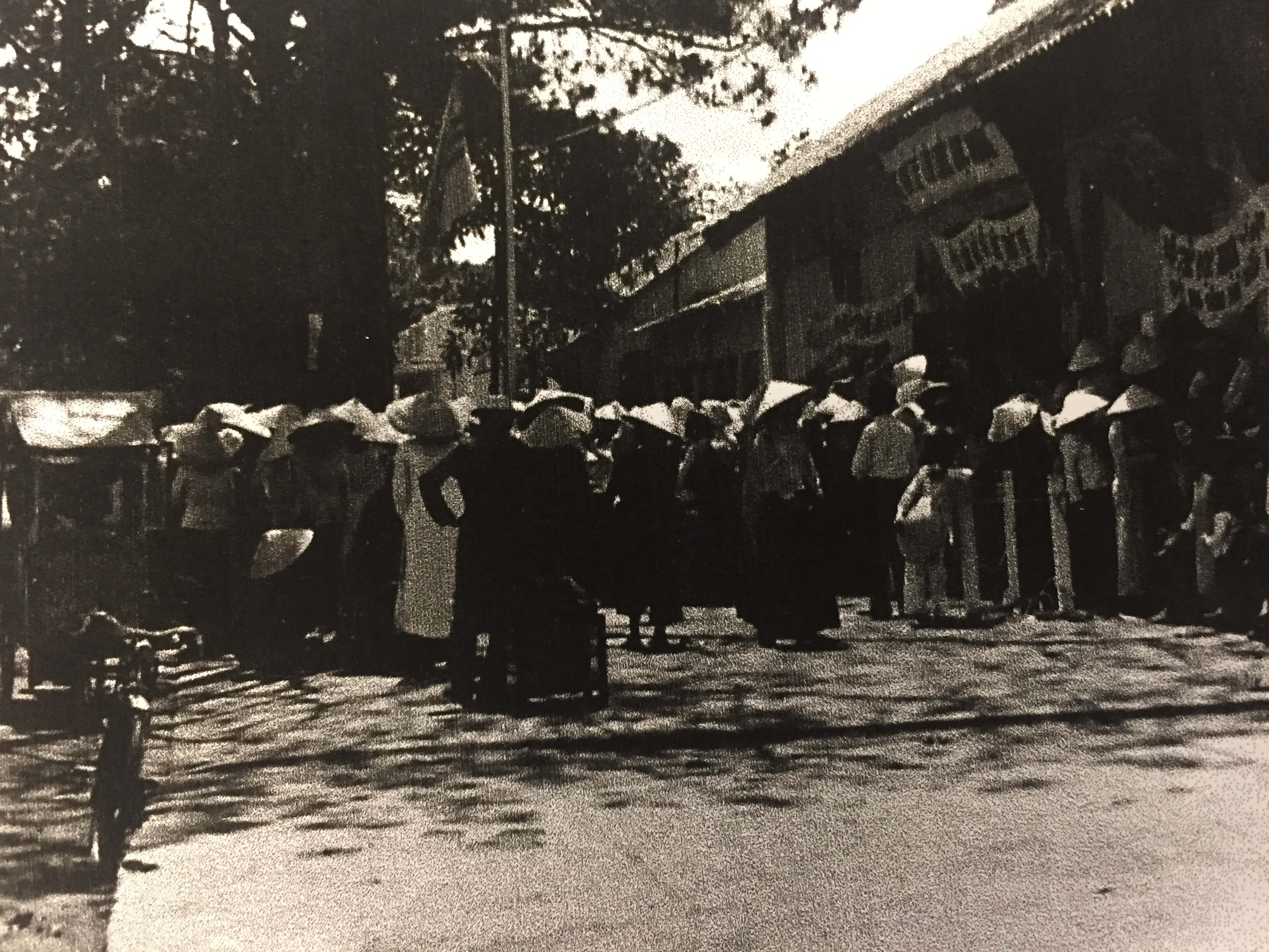 Women in straw, conical hats gathered in the dappled shade of the streets of a town.