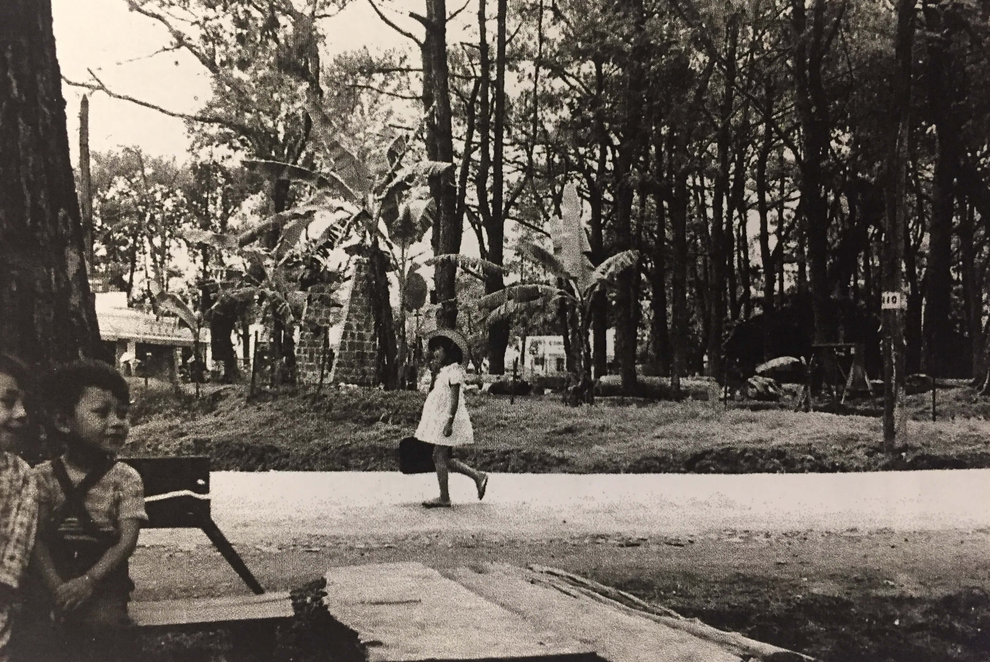 Young Asian child in white dress walking along the road. Two very young boys on a bench in the left foreground.