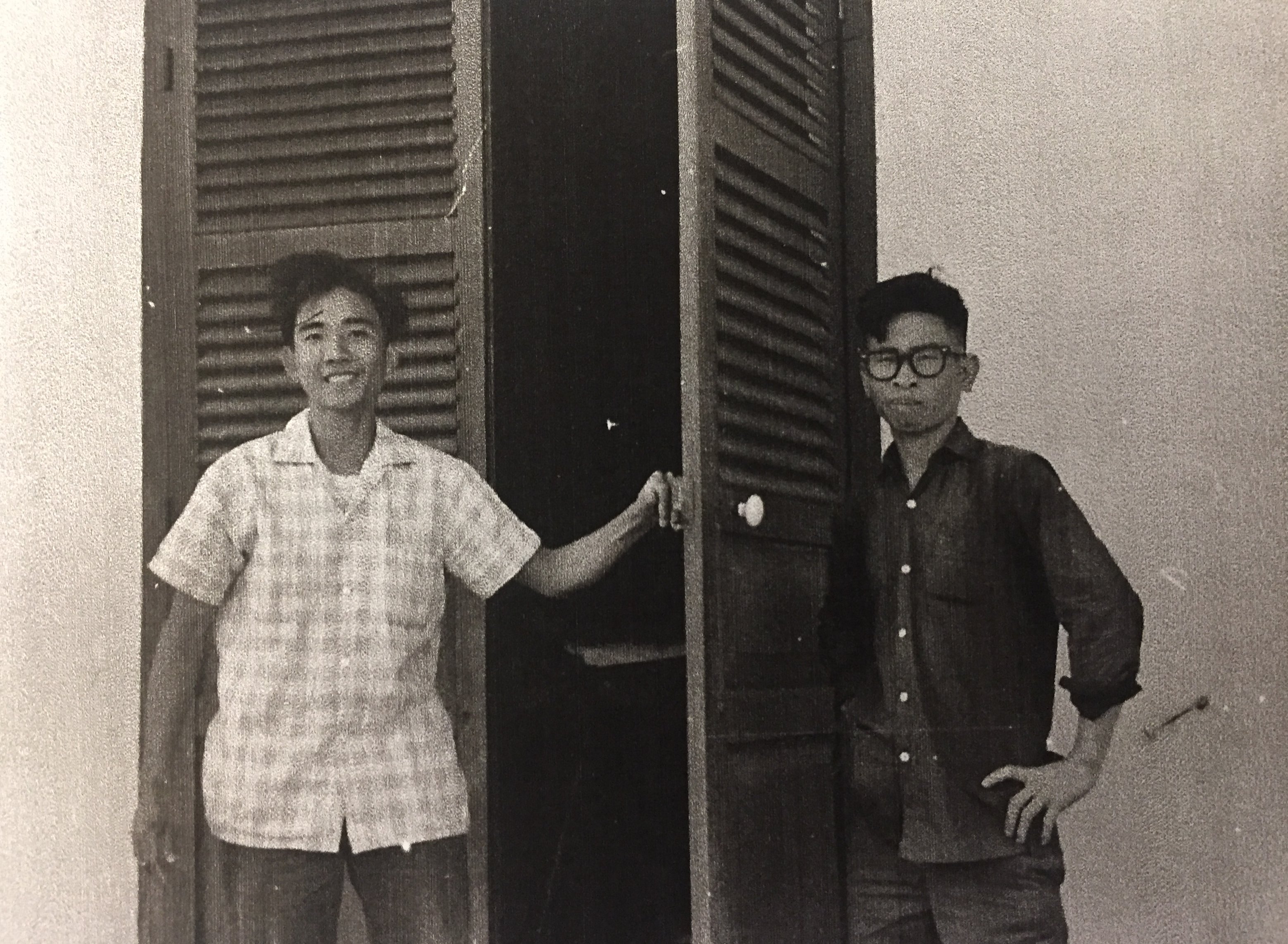 Two young Asian men standing outside a building with shutter doors. One wears a light colored plaid shirt, the other is in black.